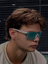 A man wearing a beige graphic t-shirt, black shorts, and blue-tinted sunglasses on an indoor tennis court. Vyzr glasses
