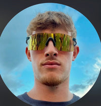 A close-up portrait of a man wearing oversized shield-style sunglasses with reflective gold mirrored lenses and black frames, set against a circular background with a blue sky and clouds behind him.