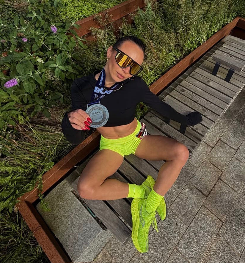 A woman in athletic wear sits on a wooden bench in an outdoor garden setting. She is wearing a black long-sleeve crop top, neon yellow shorts, neon yellow socks, and reflective gold sunglasses, holding a small round tin.