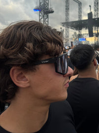 A person in a crowd at an outdoor concert or event, wearing black sunglasses with a visible VFT brand mark on the temple, against a backdrop of stage lighting towers and a cloudy sky.