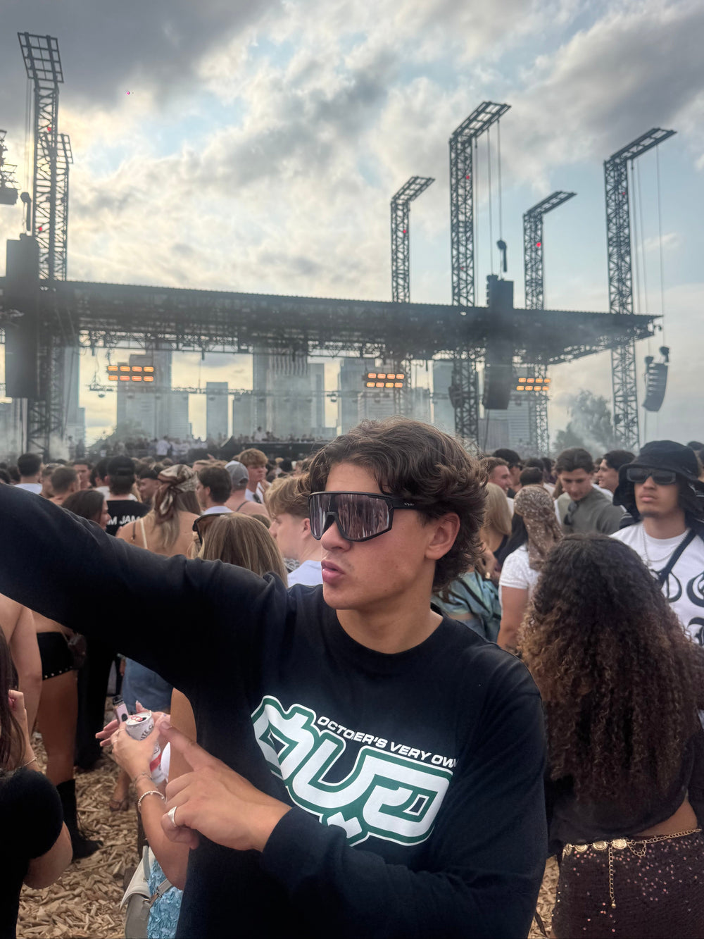 A person at an outdoor concert wearing a black long-sleeve shirt with a green and white graphic that reads 'OUTSTANDING VERY COOL' and 'GOD', standing in front of a large stage and cloudy sky. wearing vyzr glasses