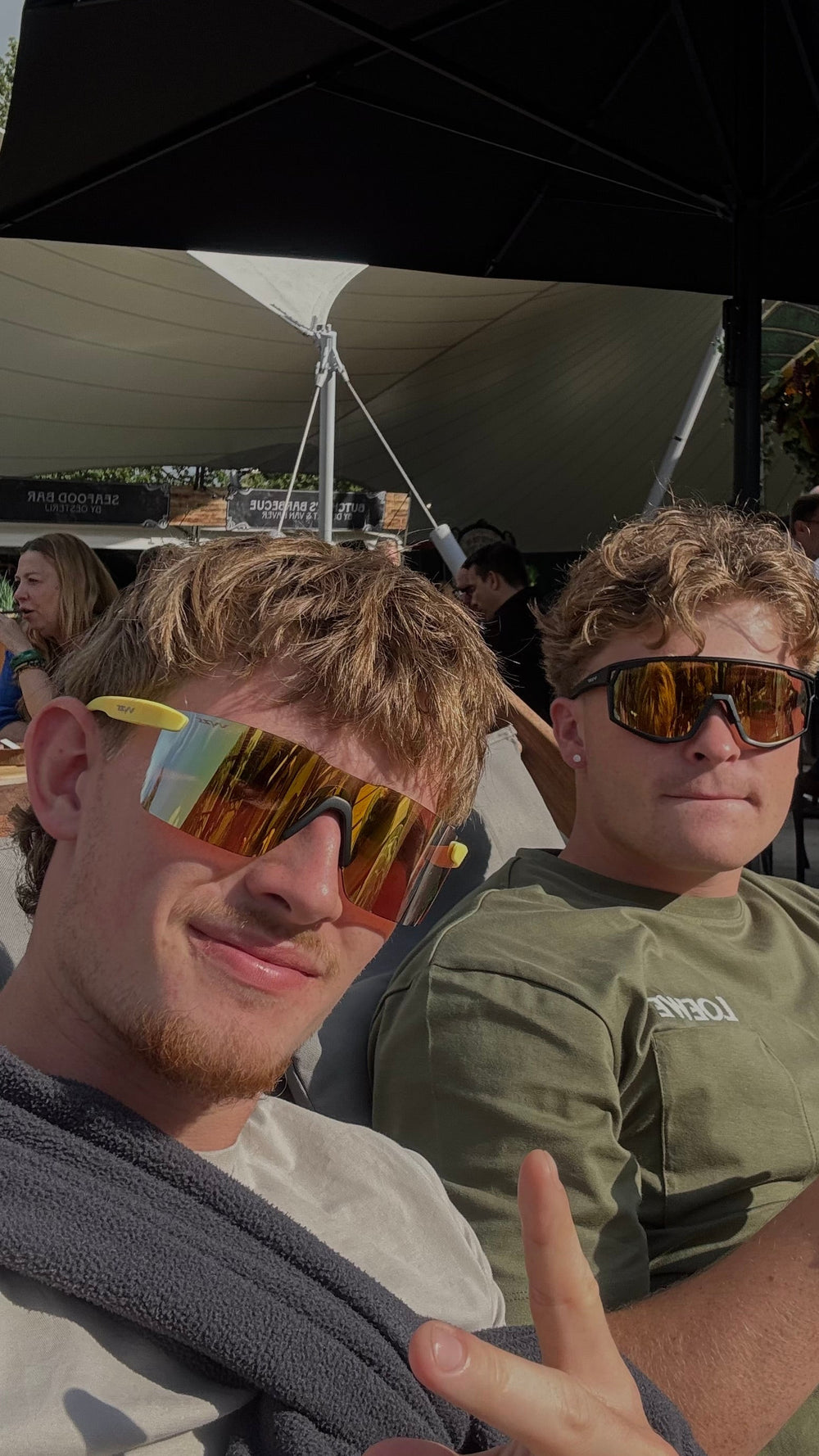 Two men wearing reflective mirrored vyzr sunglasses sitting under a large black patio umbrella at an outdoor event. The man on the left is taking the photo and wearing a gray towel over his shoulder, while the man on the right wears an olive green t-shirt with a visible 'NBJ' logo.