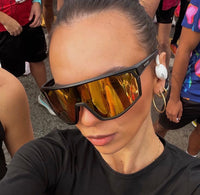 A close-up selfie of a woman wearing black athletic sunglasses with reflective gold mirrored lenses, a black long-sleeve crop top, and white earbuds. She is outdoors at what appears to be a race or event, with other participants visible in the background.