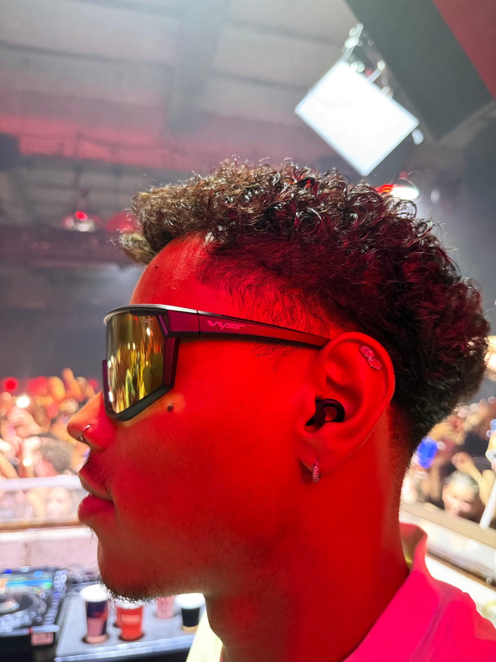 A close-up side profile of warehouse project DJ wearing Vyzr black wrap-style sunglasses with reflective gold mirrored lenses, set against a red-lit concert or club background.
