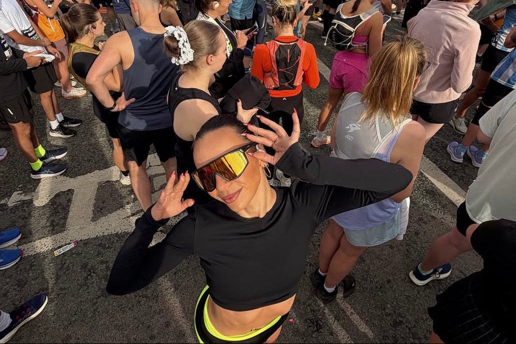 A large crowd of people gathered outdoors on a street or parking area at sunset, with a woman in the foreground wearing a black long-sleeve athletic crop top, neon yellow shorts, and reflective yellow-tinted sunglasses.