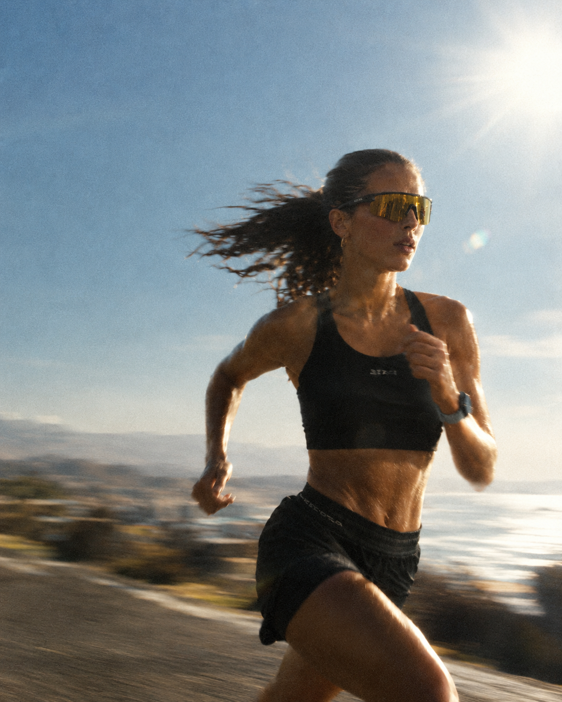A woman running outdoors wearing a black sports bra and black shorts, with yellow-tinted sunglasses and a smartwatch. The background shows a blurred coastal landscape under a bright sun and blue sky.